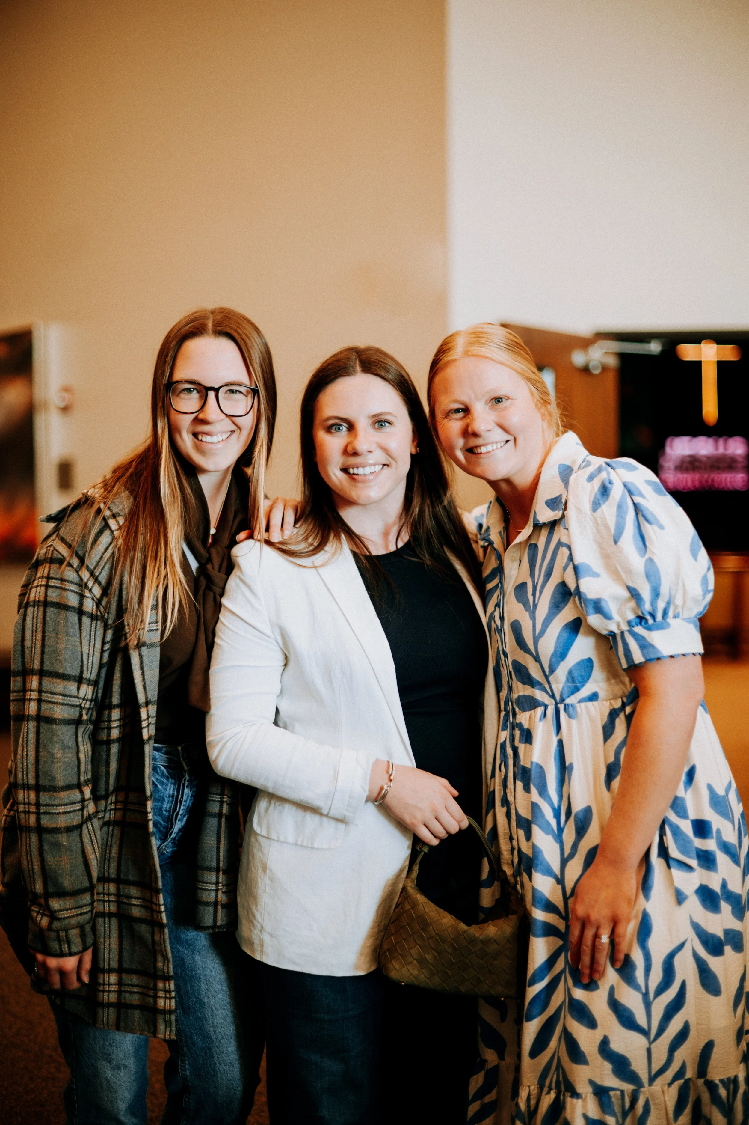 Three women smiling and standing close together indoors, with one woman wearing glasses and a plaid jacket, another in a white blazer, and the third in a light blue and white patterned dress.