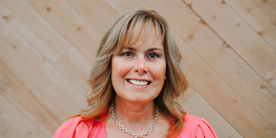 A woman with shoulder-length blonde hair, smiling, wearing a pink top and a silver chain necklace, standing in front of a wood-paneled wall.