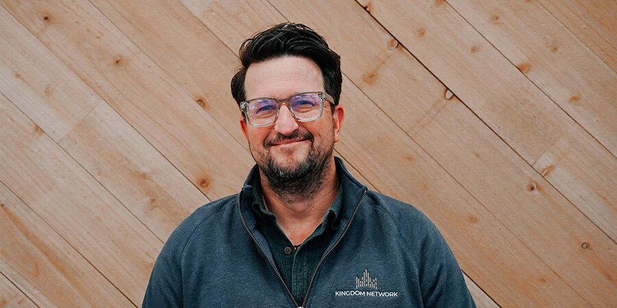 A smiling man with dark hair and glasses, wearing a dark zip-up jacket with a logo, standing in front of a wood-paneled wall.