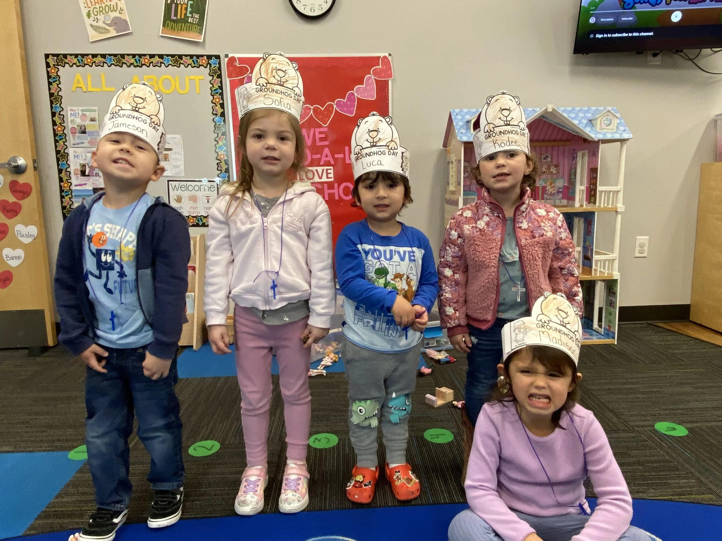 Five young children standing in a classroom, wearing paper hats with a Groundhog Day theme, inside a classroom with colorful posters and toys.