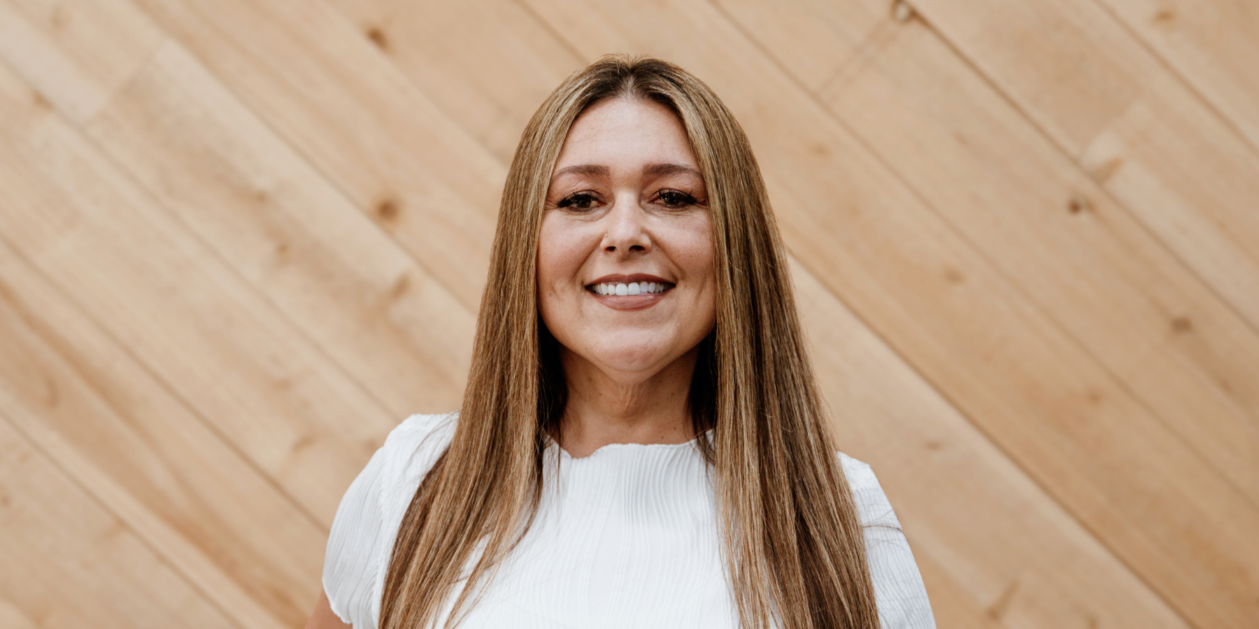 A woman with long straight hair, smiling, wearing a white top, standing in front of a wooden wall