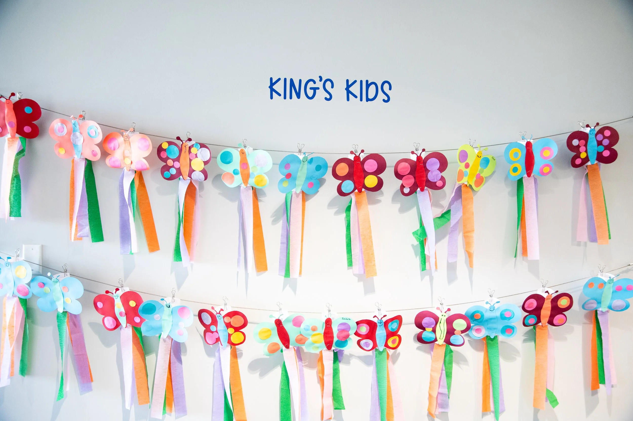 Colorful butterfly decorations with paper wings and ribbon bodies hanging on a string, along with a sign that says 'KING'S KIDS' on a white wall.