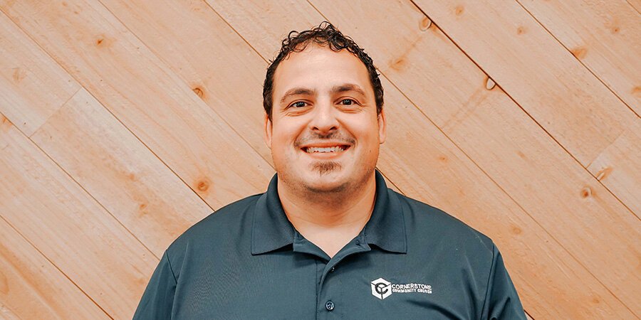 A smiling man wearing a dark polo shirt with a 'Cornerstone Community Church' logo stands in front of a light wood-paneled wall.