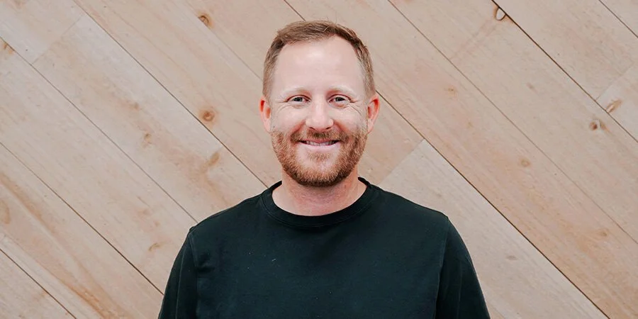 Smiling man with short hair and beard wearing a black shirt standing in front of a wooden wall.