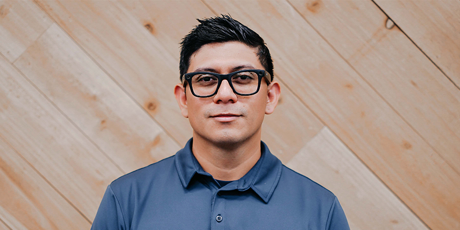 Portrait of a man wearing glasses and a blue collared shirt standing against a wooden wall background.