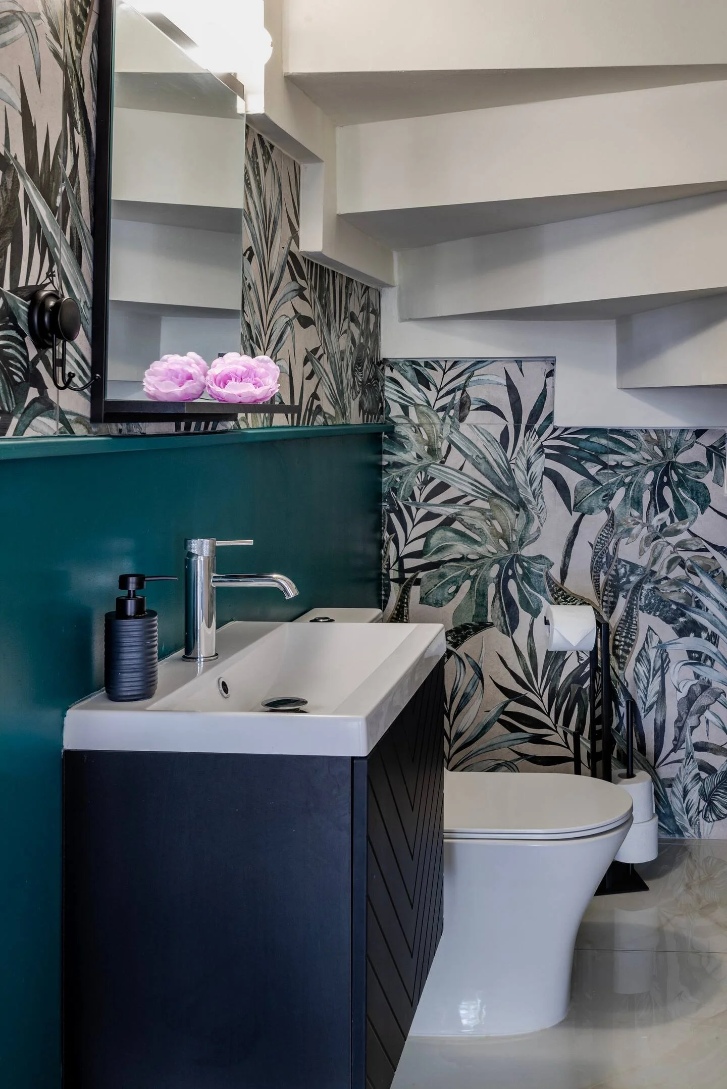 Modern bathroom with dark vanity, white sink, black soap dispenser, large mirror, botanical wallpaper, white toilet, toilet paper holder, and decorative flowers.