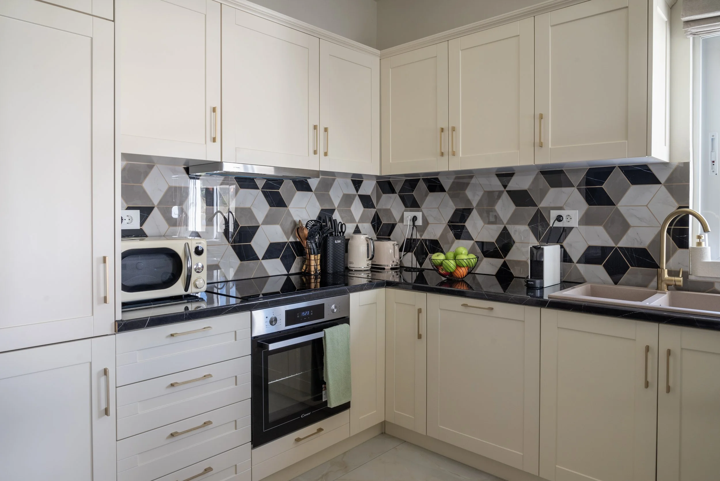 Modern kitchen with white cabinets, black and beige geometric backsplash, black countertop, and various small appliances, including a microwave, toaster, and a bowl of green apples.