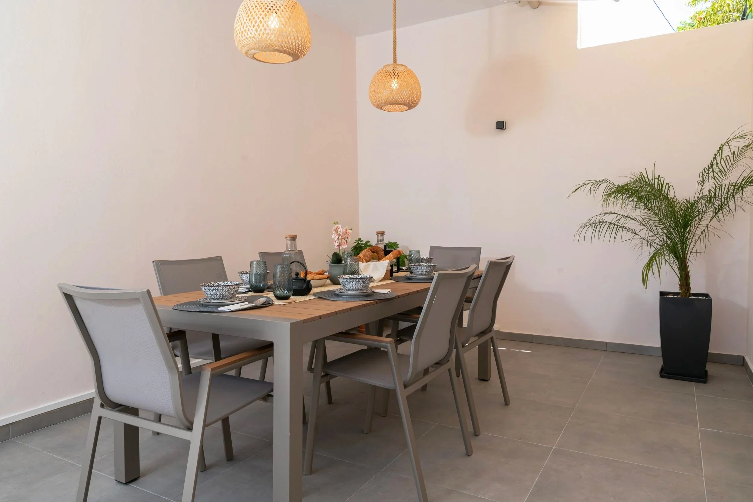 Modern dining room with a rectangular wooden table set with bowls, glasses, and a centerpiece, six light gray chairs, two wicker pendant lights, large potted plant in a black square planter, and a small window on the wall with a view of greenery.