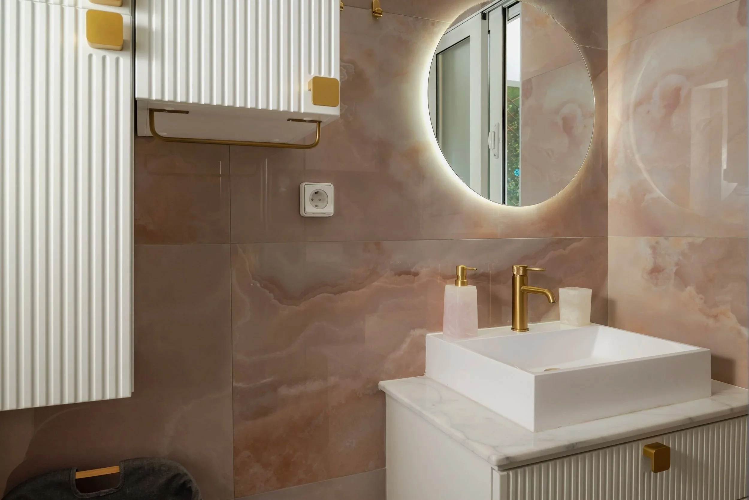 Modern bathroom with a marble countertop sink, gold faucet, circular backlit mirror, pink soap dispensers, and wall-mounted radiator in the background.