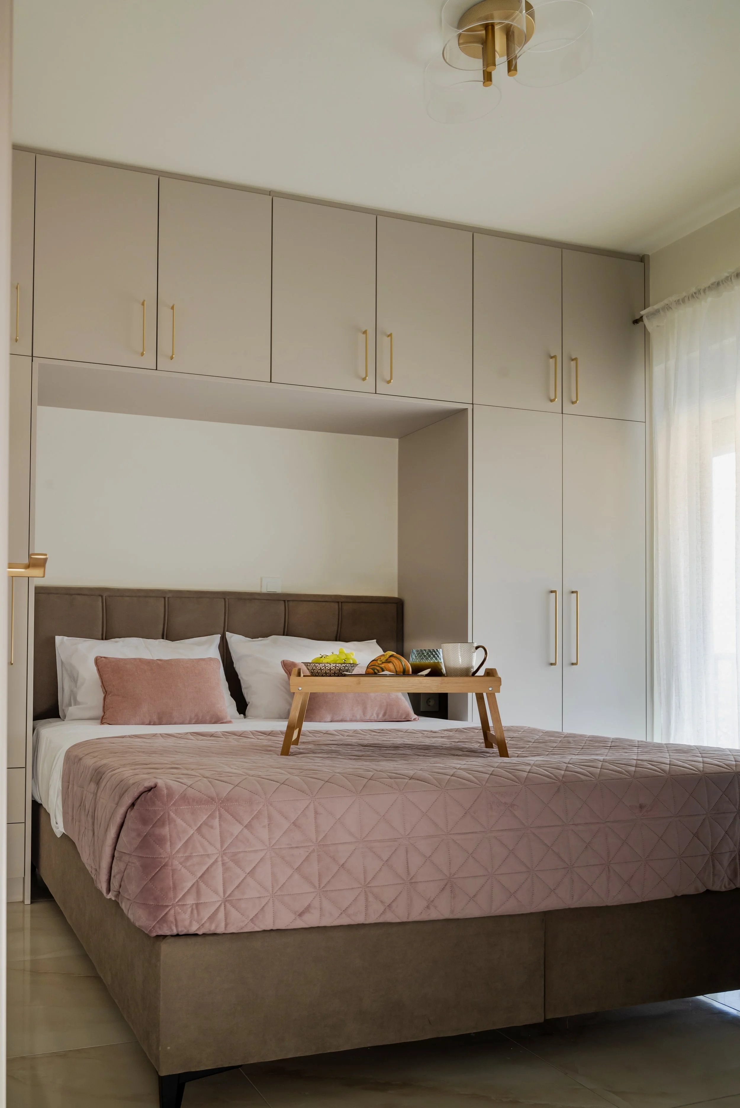 A bedroom with a large bed that has a pink quilt and two matching pillows. A tray with breakfast items, including a teapot and a cup, sits on the bed. There are built-in cabinets with gold handles above the bed and a curtain on the window to the righ