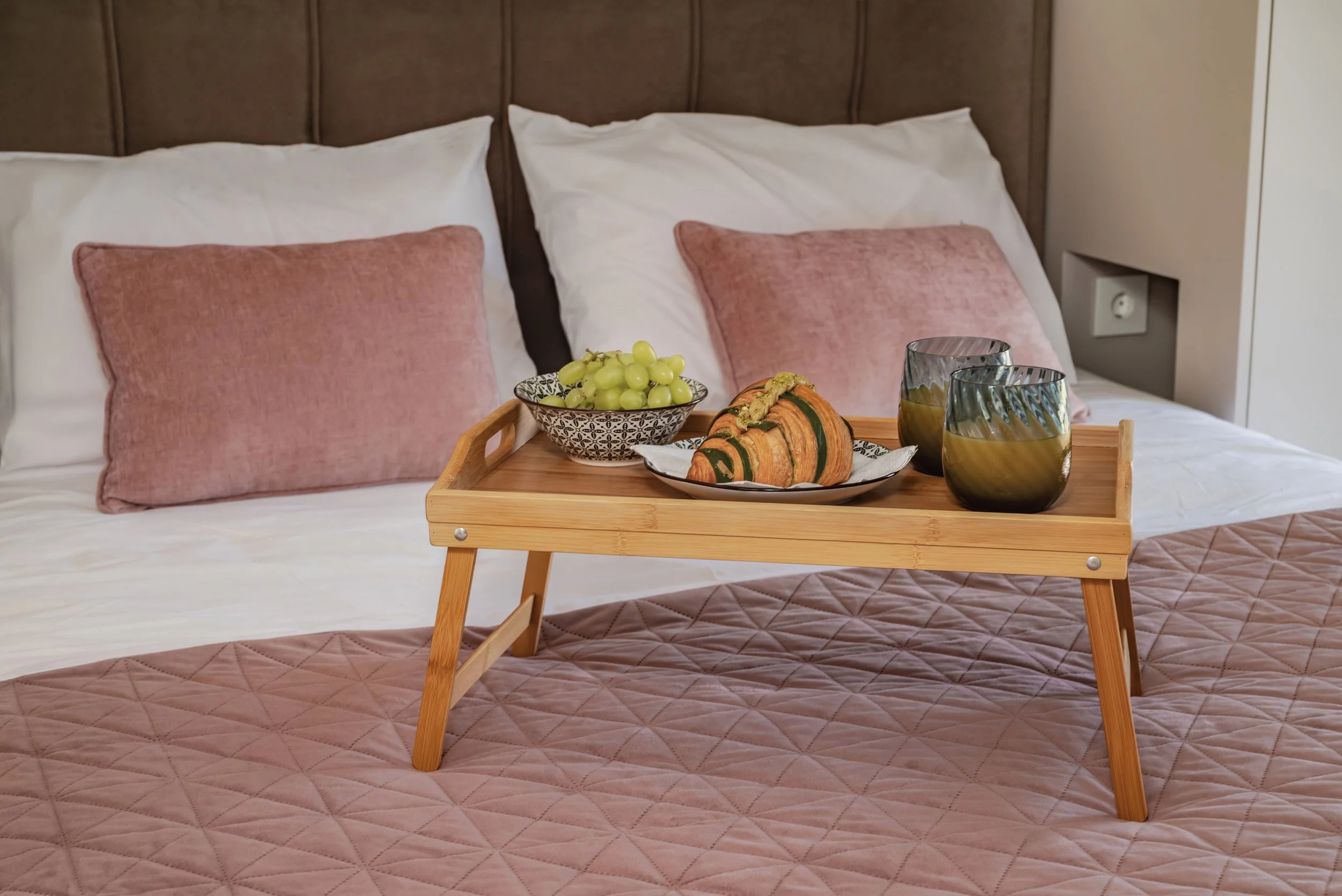 A bed with pink pillows and quilted bedspread, a wooden tray with grapes, pumpkin, and two glasses on it.