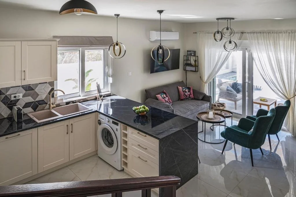 Open-concept kitchen and living room with kitchen featuring white cabinets, black marble countertop, and pink sink; living area with gray sofa, two green armchairs, wall-mounted TV, and sliding glass door with white curtains leading to a balcony.