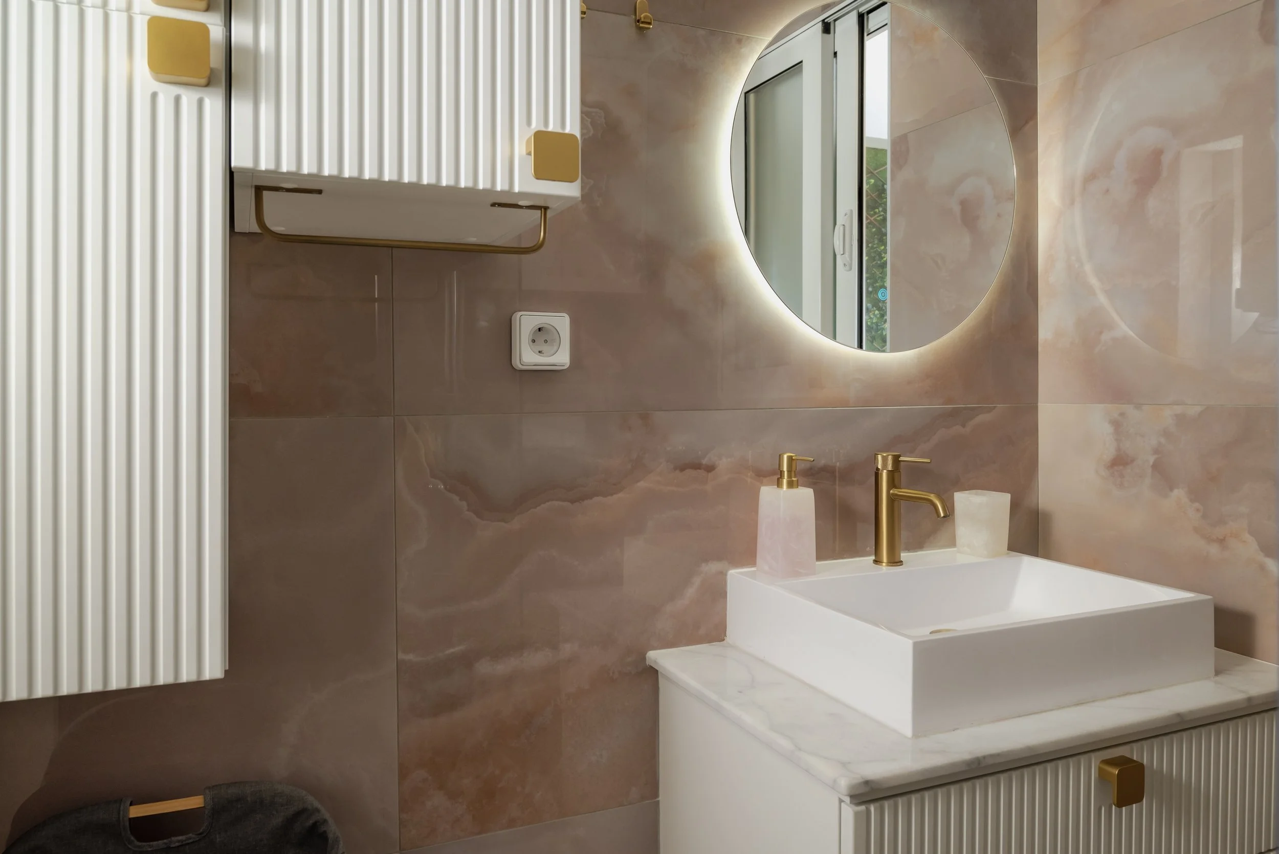 Modern bathroom with pink marble walls, a round backlit mirror, a white vessel sink with gold fixtures, and a window reflecting greenery outside.
