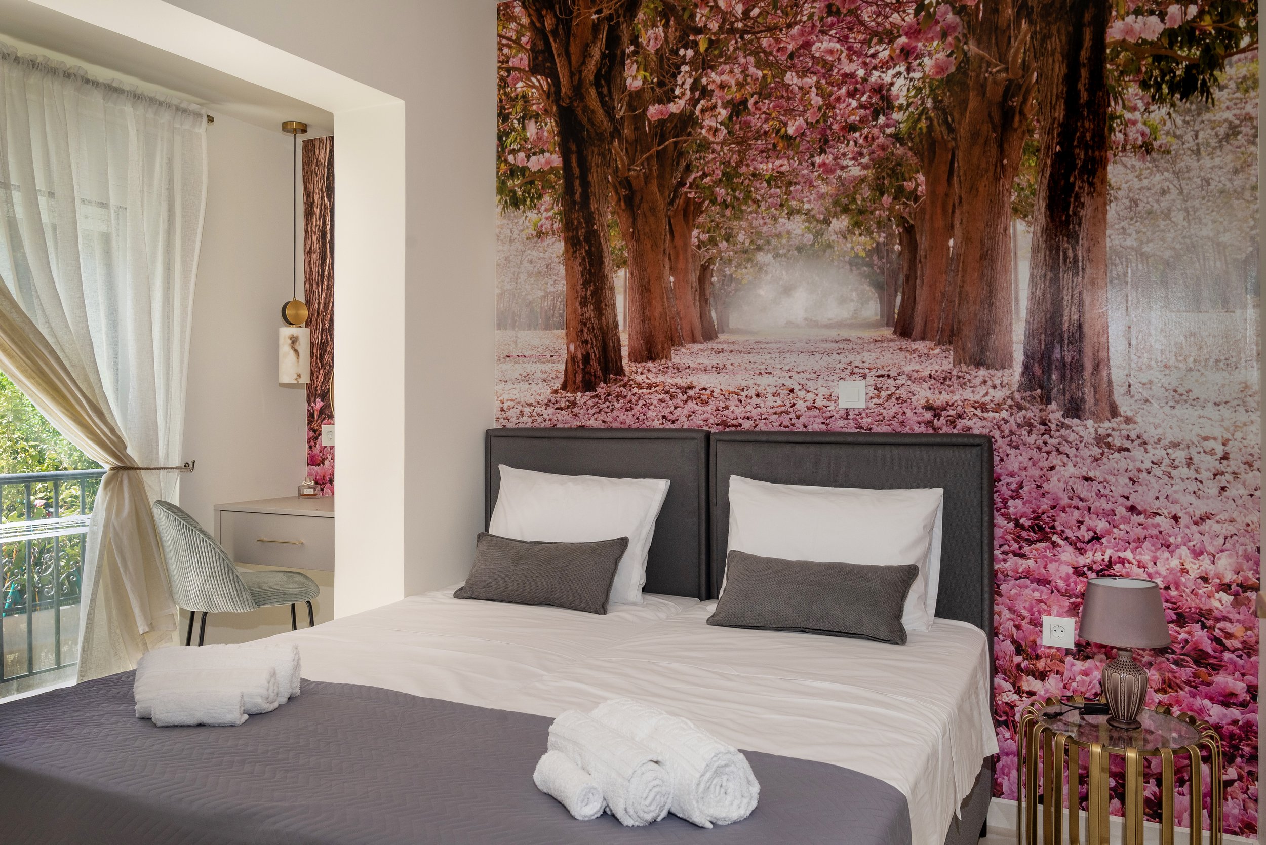 Bedroom with large wall mural of pink flowering trees on a pathway, a bed with gray headboard and white bedding, towels on the bed, and a small side table with a lamp.