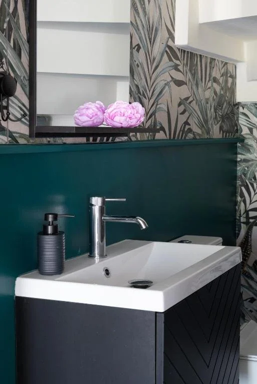 Bathroom sink with a silver faucet and a black soap dispenser, with decorative pink bath poufs and leafy wallpaper in the background.