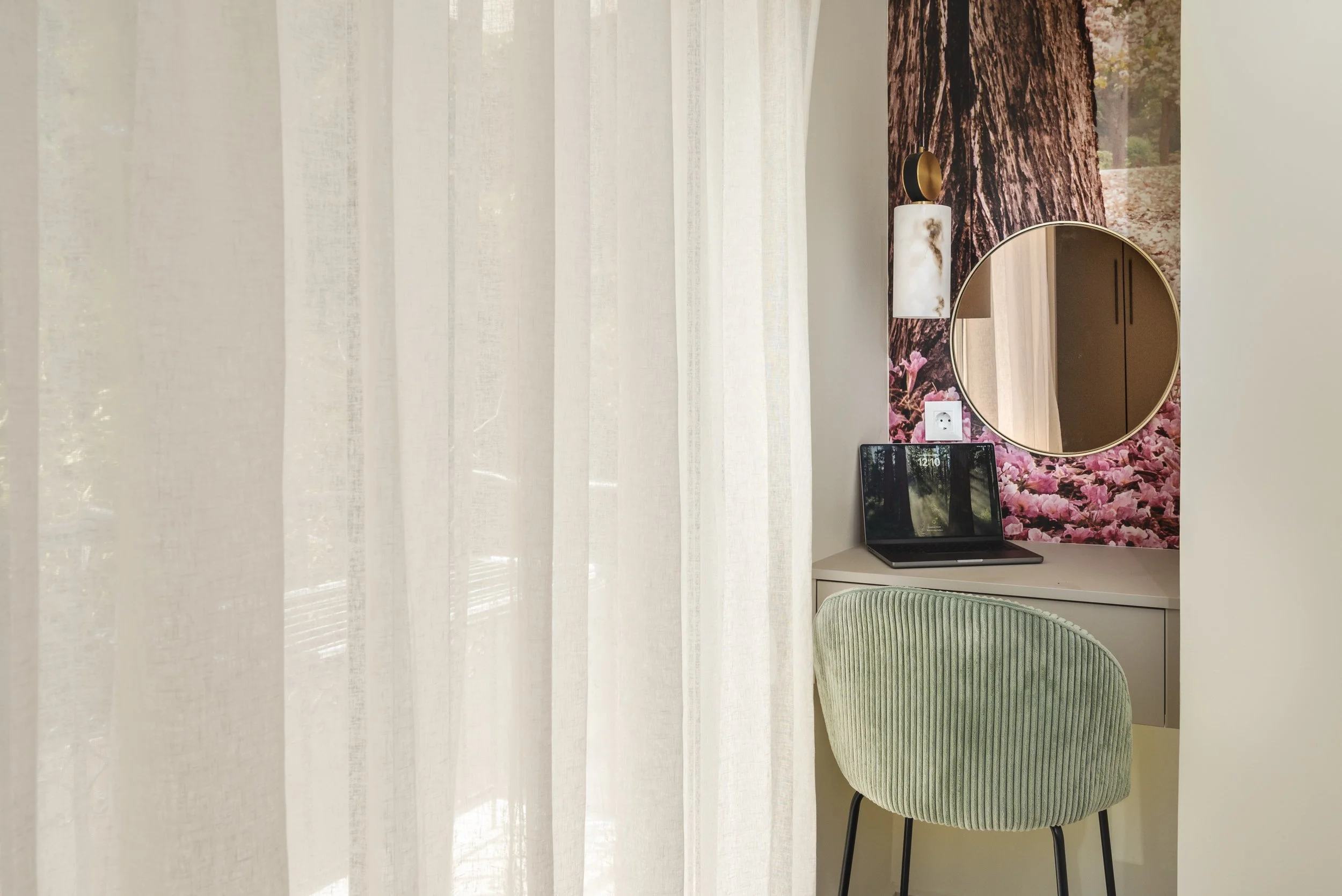 A small workspace with a desk, a green upholstered chair, a laptop, a round mirror, and wall art of a large tree trunk with pink flowers in the background.