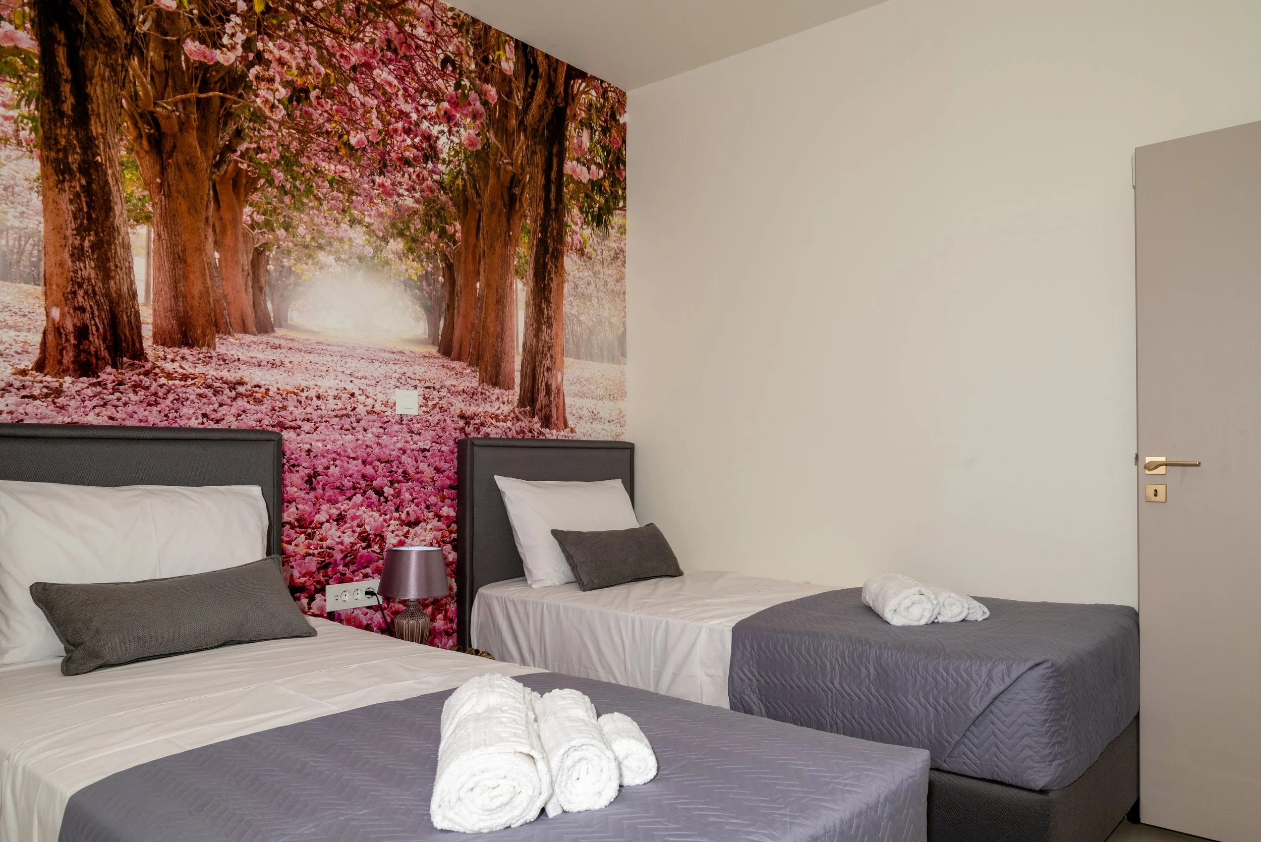 A bedroom with two beds, each with white and gray bedding and rolled white towels on top. Behind the beds is a large wall mural depicting a pink flowering tree-lined path with sunlight filtering through the branches.