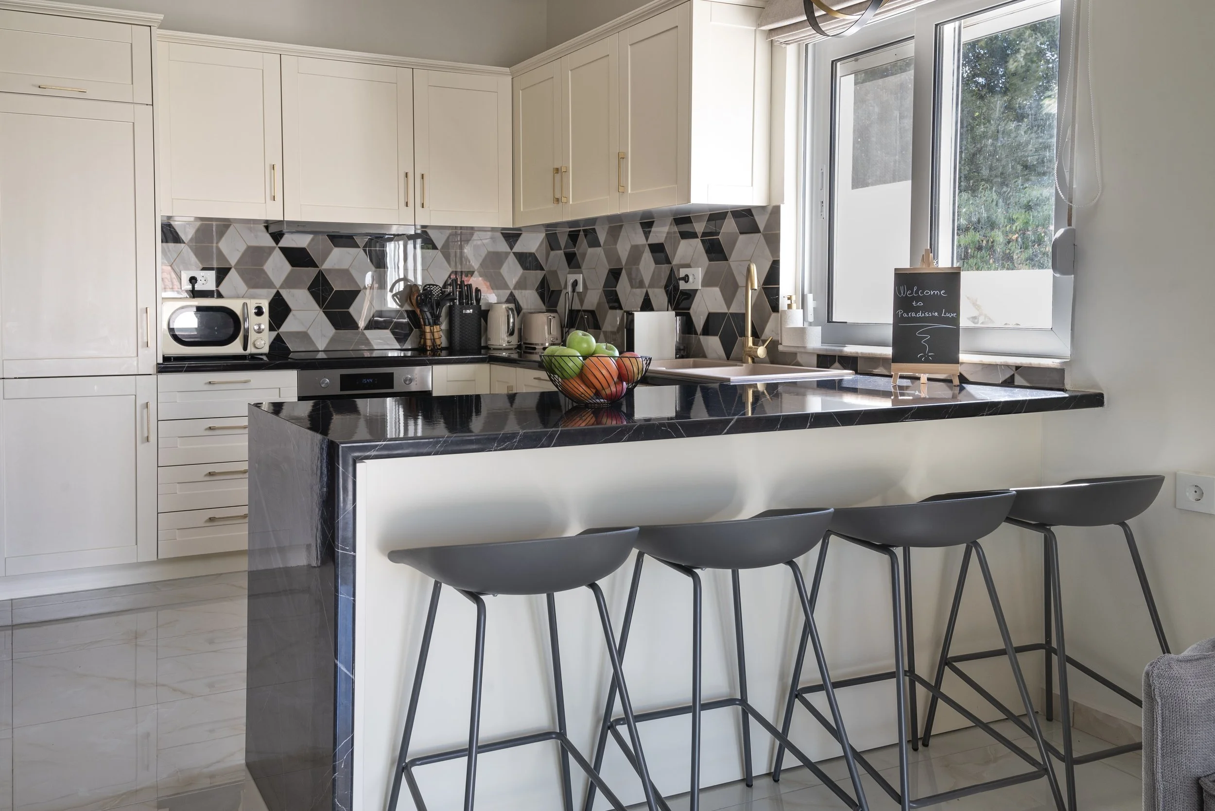 Modern kitchen with white cabinets, black countertops, and geometric patterned tile backsplash. There are four black barstools at the counter, which has a bowl of apples and a chalkboard sign near the window.