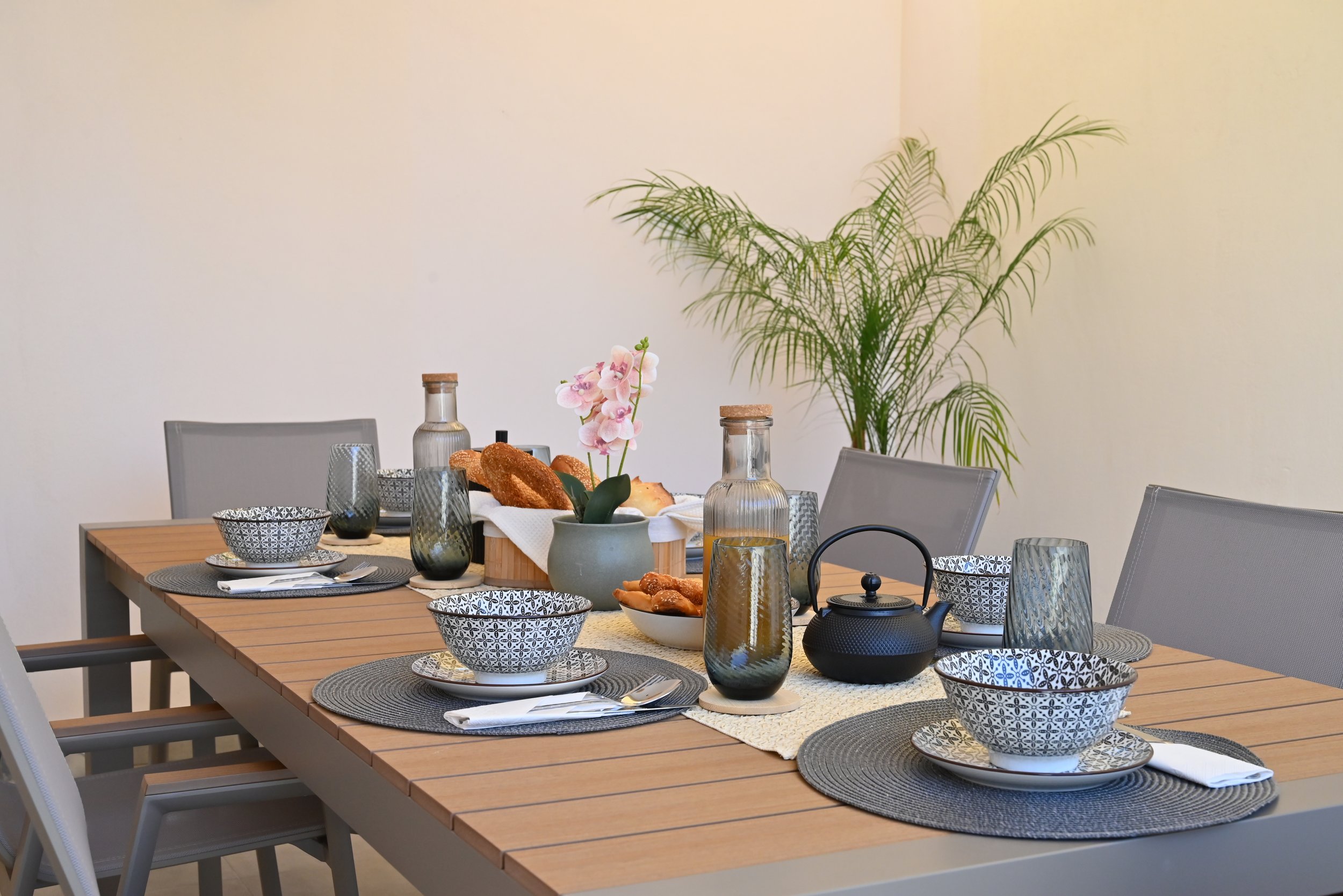 A dining table set for a meal with patterned bowls and plates, glasses, a teapot, a cream-colored table runner, and a centerpiece with a pink orchid, bread, and beverages, in a room with a large leafy plant against a plain wall.