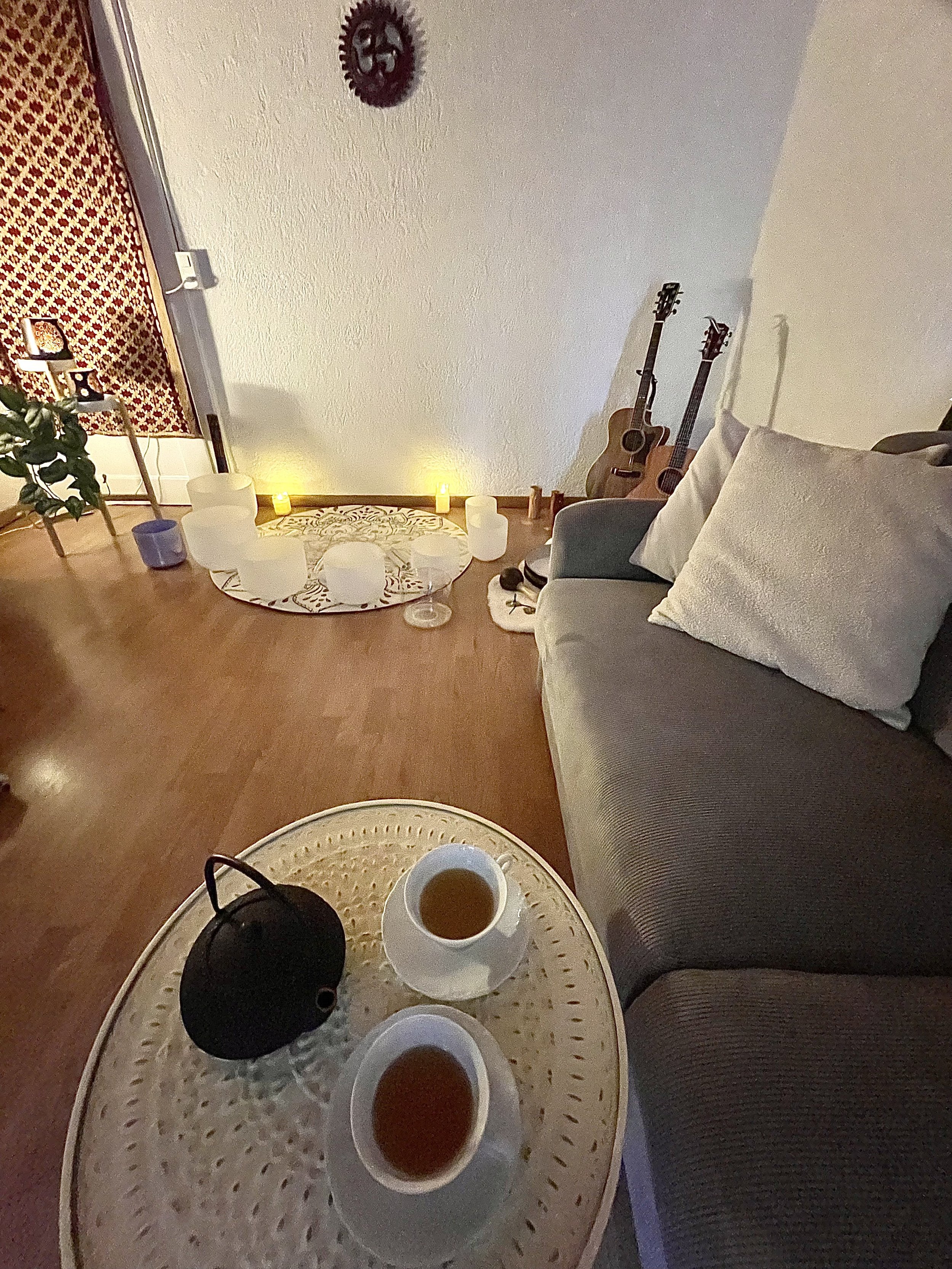 Indoor living room with a gray sofa and white pillows, two acoustic guitars leaning against the wall, lit candles on the floor, and a tea set with a kettle and three cups on a wicker table.