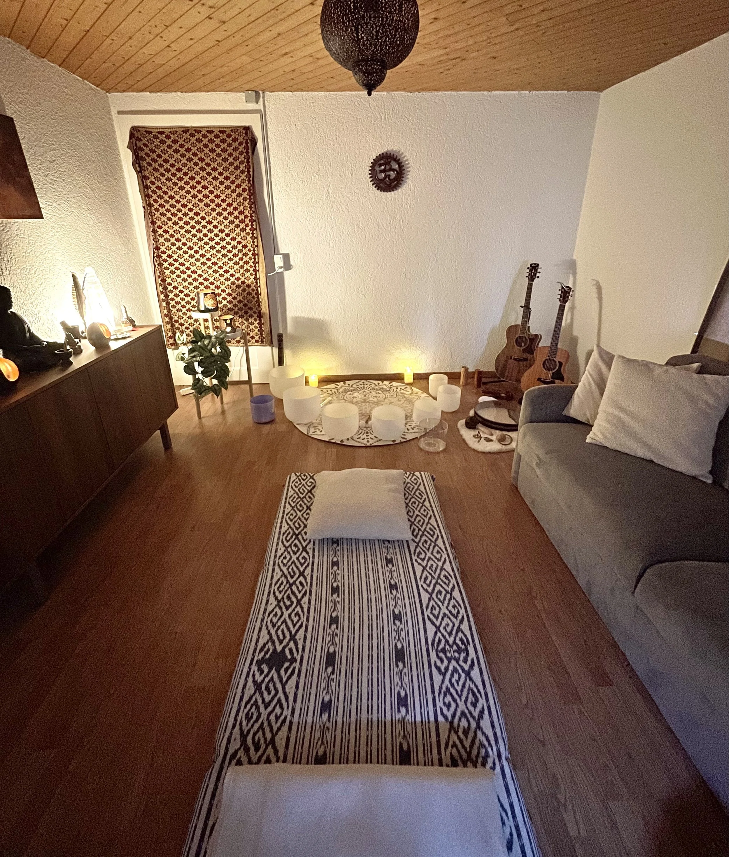 A cozy living room with a wooden sloped ceiling, textured white walls, and a patterned curtain. The room is decorated with candles, musical instruments, and potted plants, creating a warm ambiance.