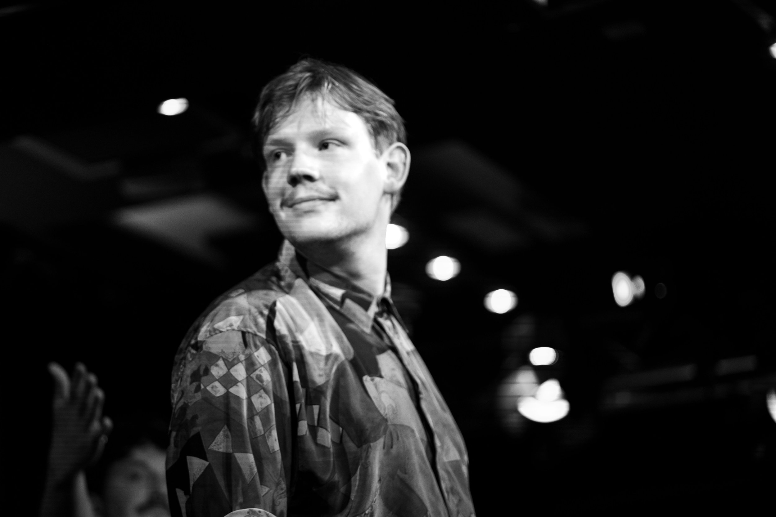 Schwarz-Weiß-Foto eines Mannes mit kurzen Haaren, der in einem dunklen Raum steht, mit Lichtern im Hintergrund, trägt ein bemustertes Hemd.