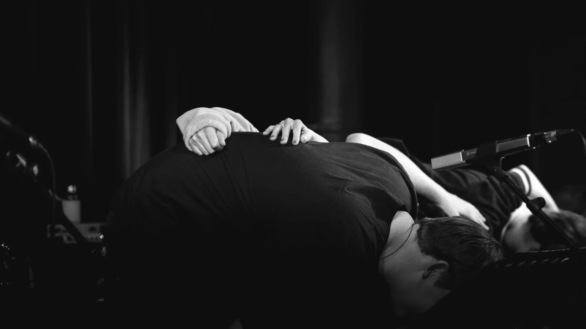 Zwei Personen liegen auf einer Bühne, eine Person hat die Hand auf den Rücken der anderen, sie scheinen zu tanzen oder zu performen.