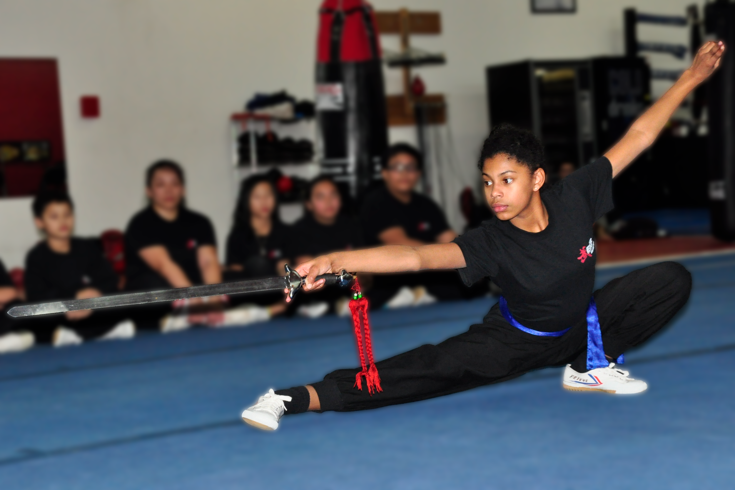 A young girl practicing martial arts, performing a high kick with a sword, in a training room with four women seated in the background watching.