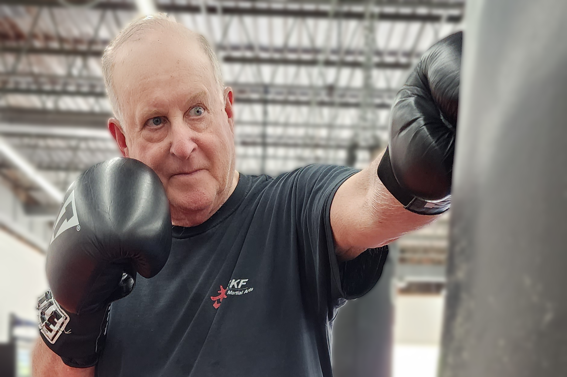 A senior man with platinum blond hair in a black T-shirt and boxing gloves practicing punching at a boxing training session in a gym.