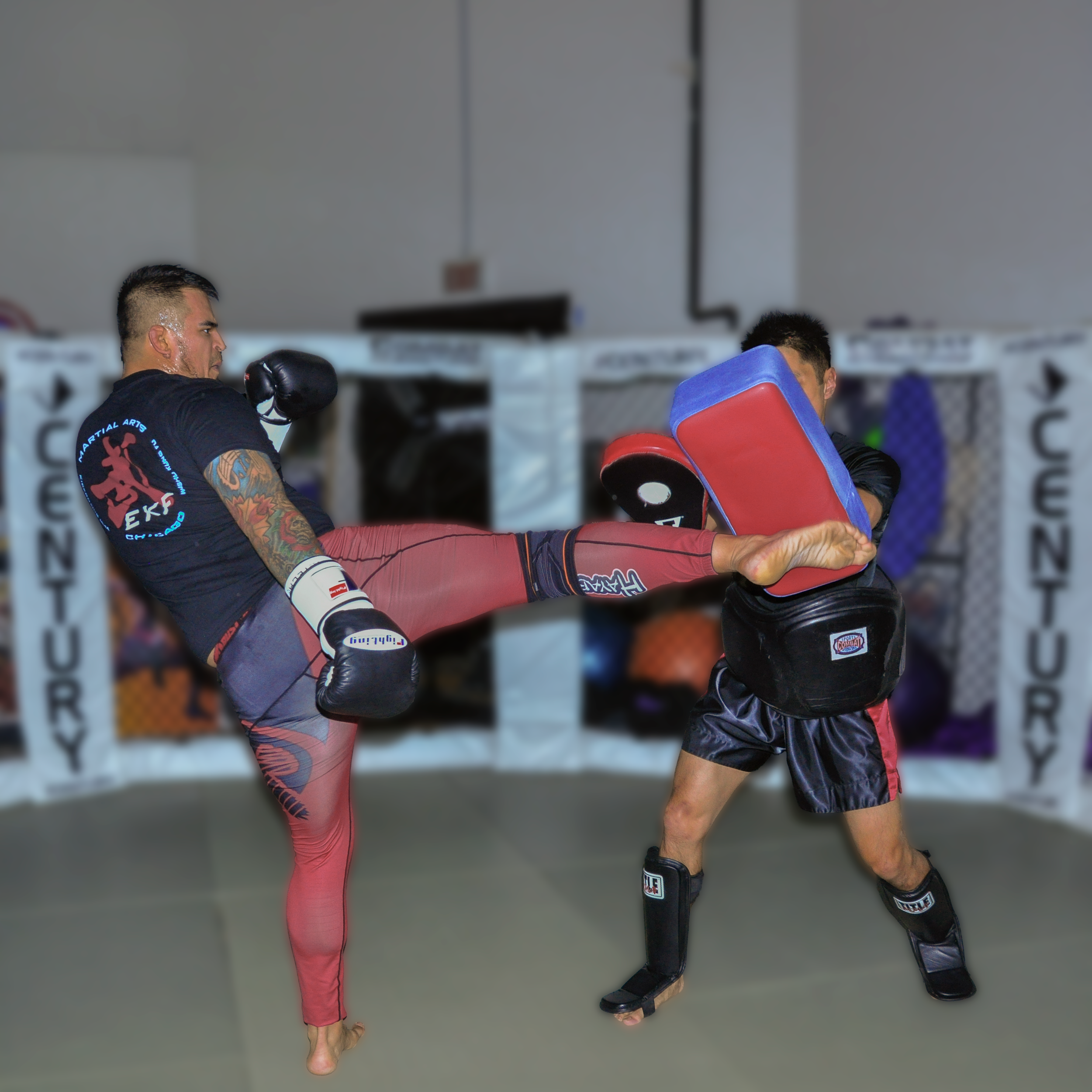 Two martial artists practicing inside a gym, one wearing red shorts and black rash guard, the other wearing black shorts and black rash guard, both in fighting stances.