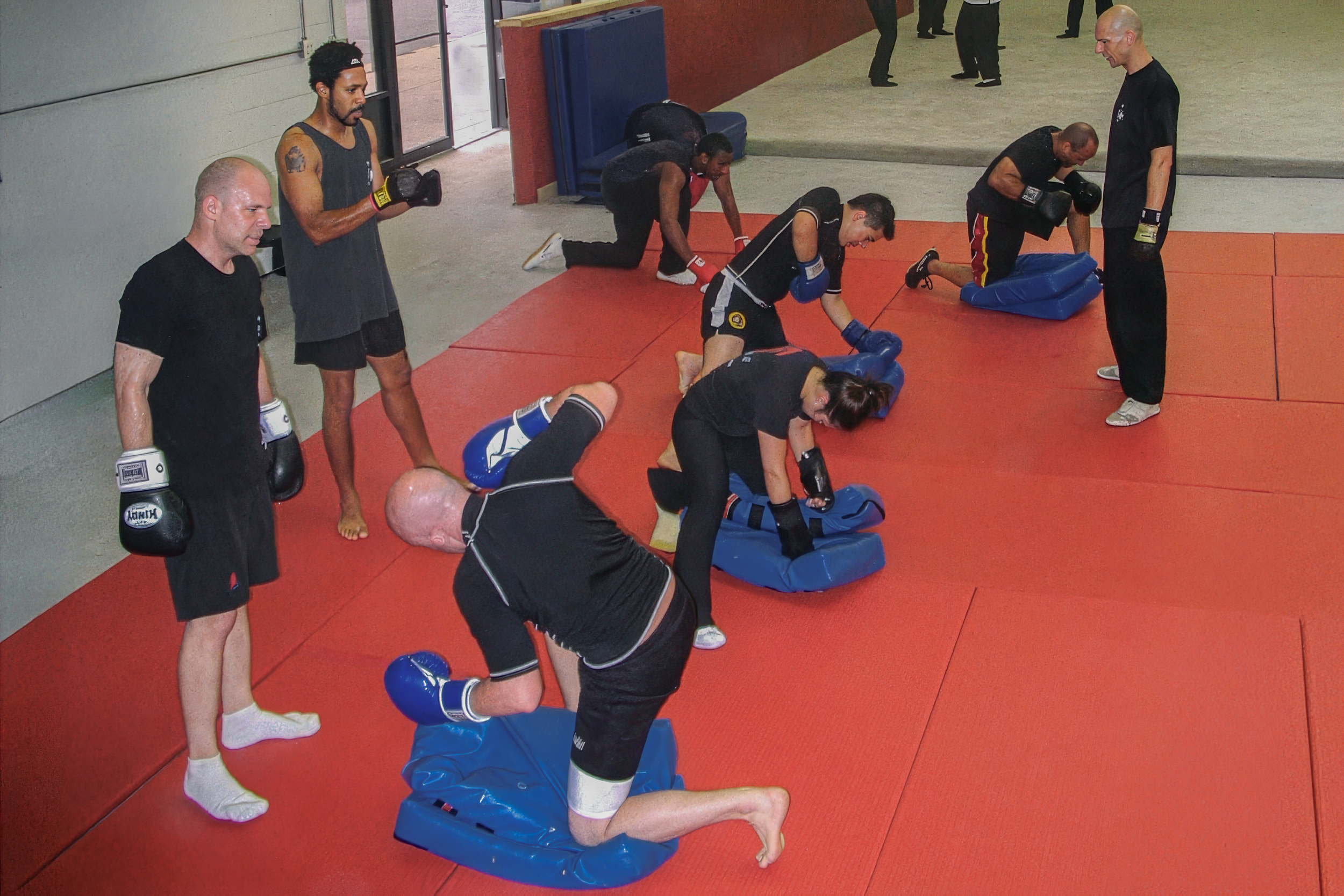 Martial arts training session with five students practicing on blue padded mats and a bald instructor overseeing. Students are in various positions, some kneeling and some on their hands and knees, wearing protective gloves and uniforms. The setting appears to be an indoor gym or dojo with a red and grey floor.