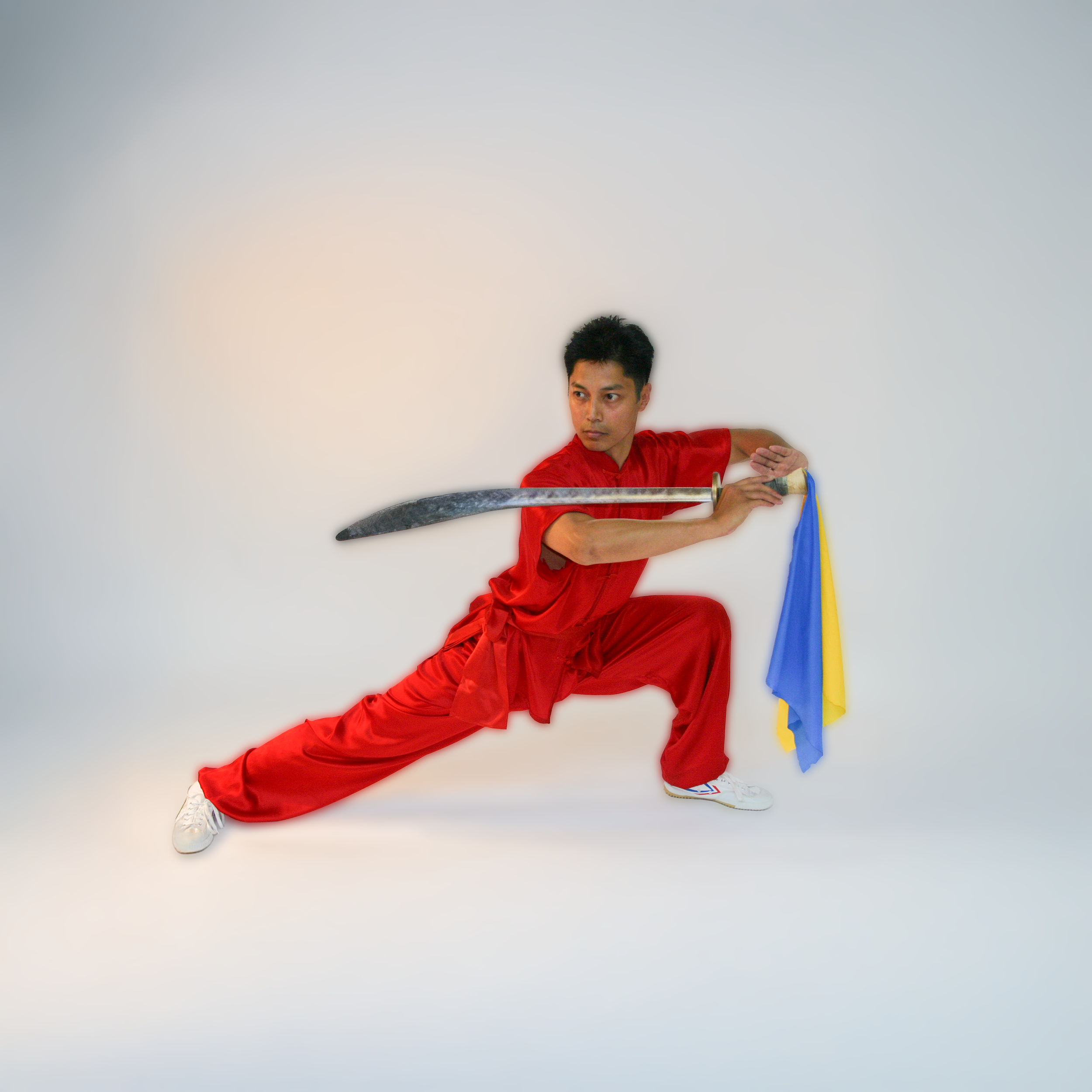 A young man in a red martial arts uniform poses in a deep stance, holding a sword with both hands, with a blue and yellow flag beside him, against a plain light background.