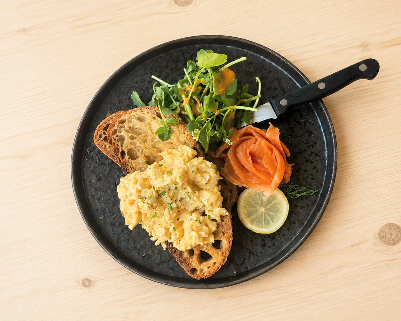 Plate with scrambled eggs on toasted bread, a slice of lemon, smoked salmon, mixed greens, and a knife, on a light wooden table.