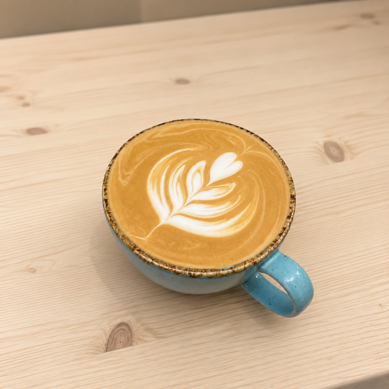 A blue ceramic mug filled with coffee topped with latte art, depicting a leaf and heart pattern, placed on a light wooden table.