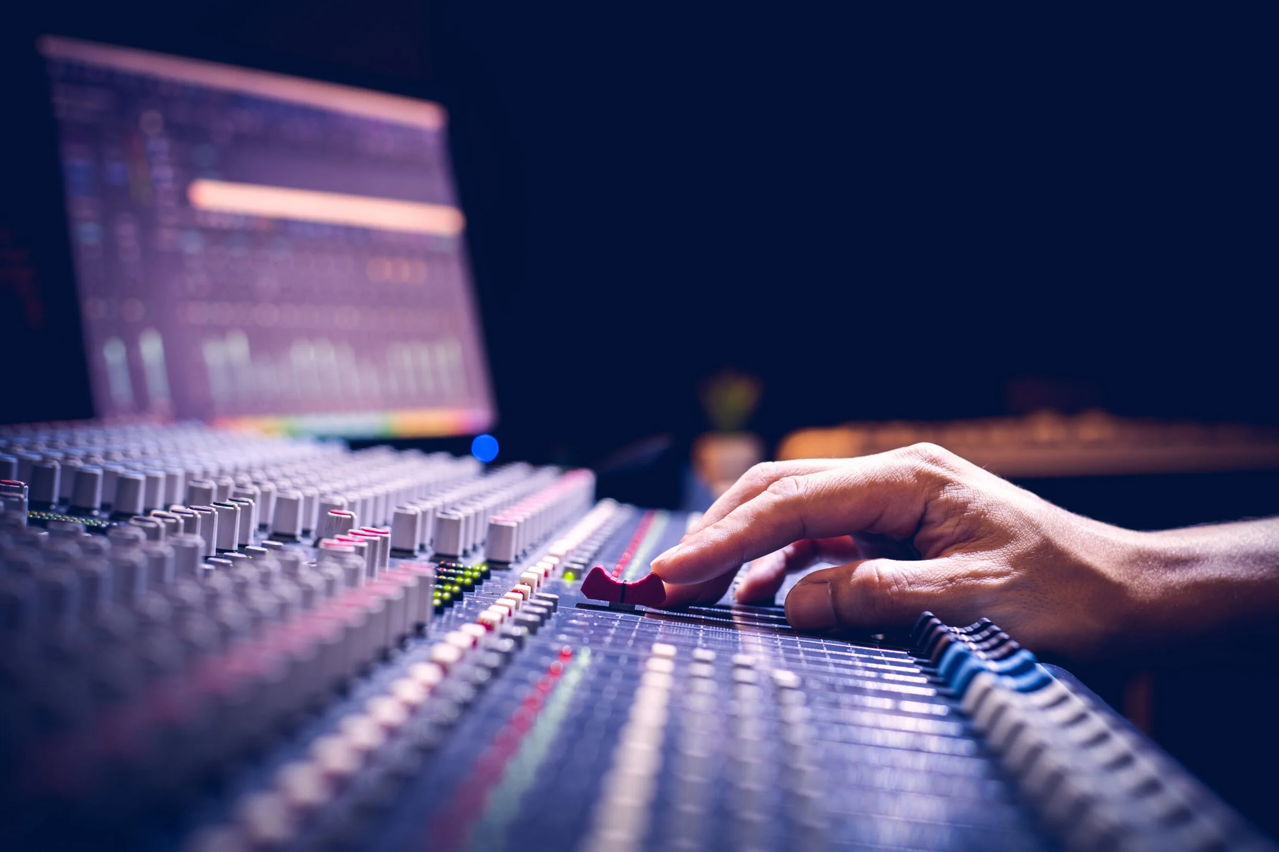 A person adjusts controls on a large audio mixing console in a dimly lit studio or control room with a computer monitor displaying audio software in the background.