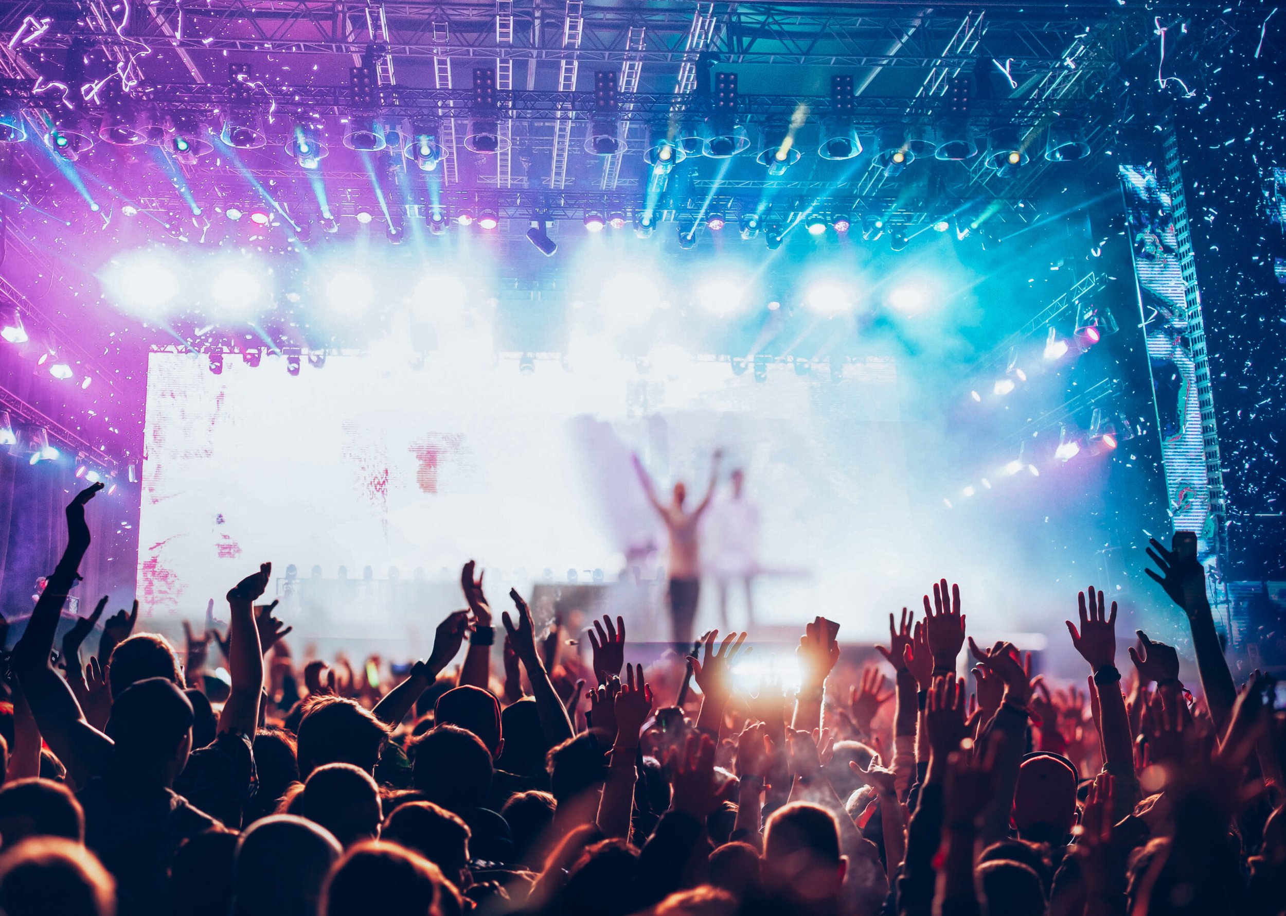 Crowd at concert with stage lights, performers, and a person with arms raised in front of bright background.