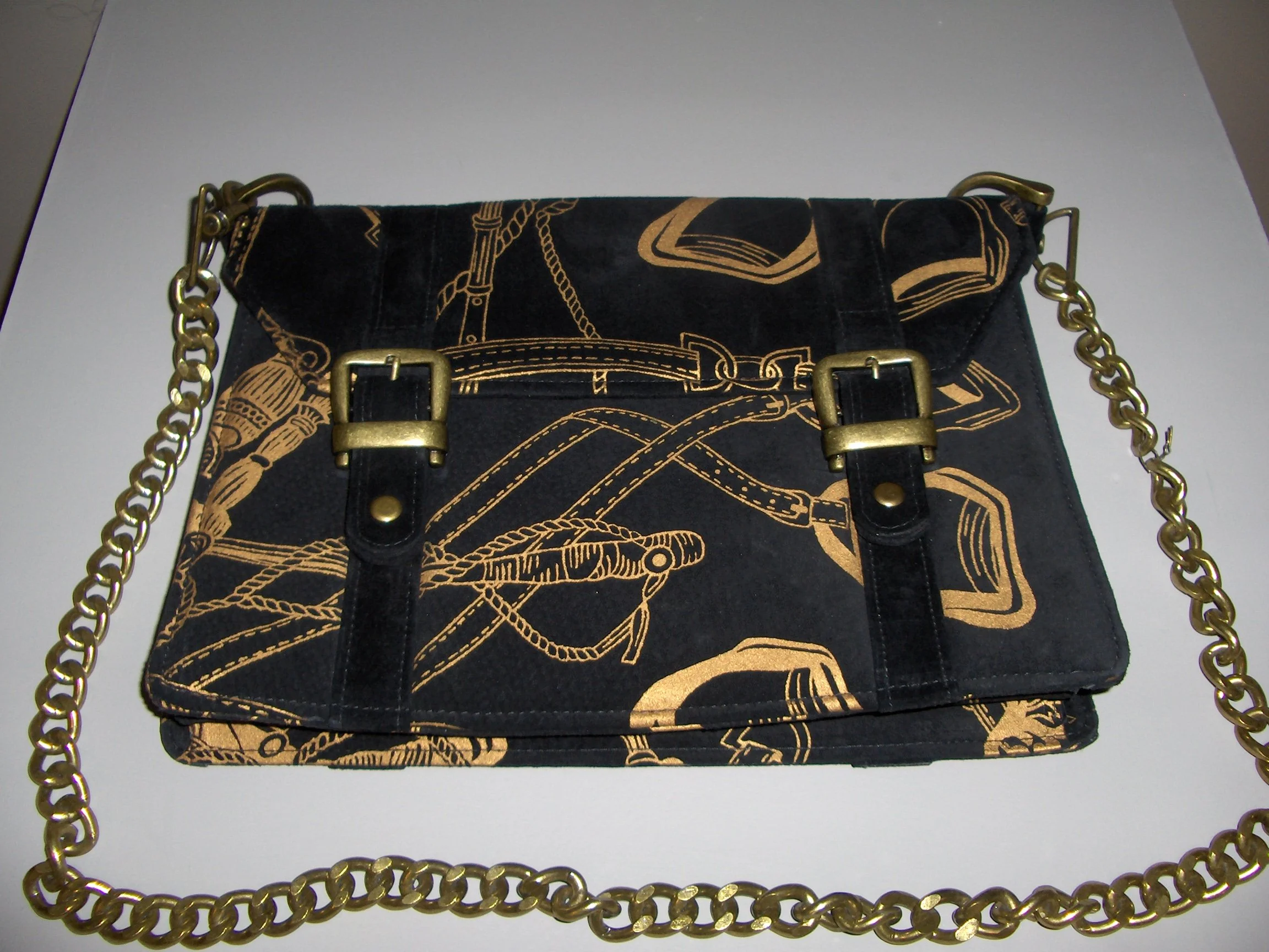 A black clutch purse with gold buckle accents and a gold chain strap, featuring a pattern of various classic artwork and design elements.