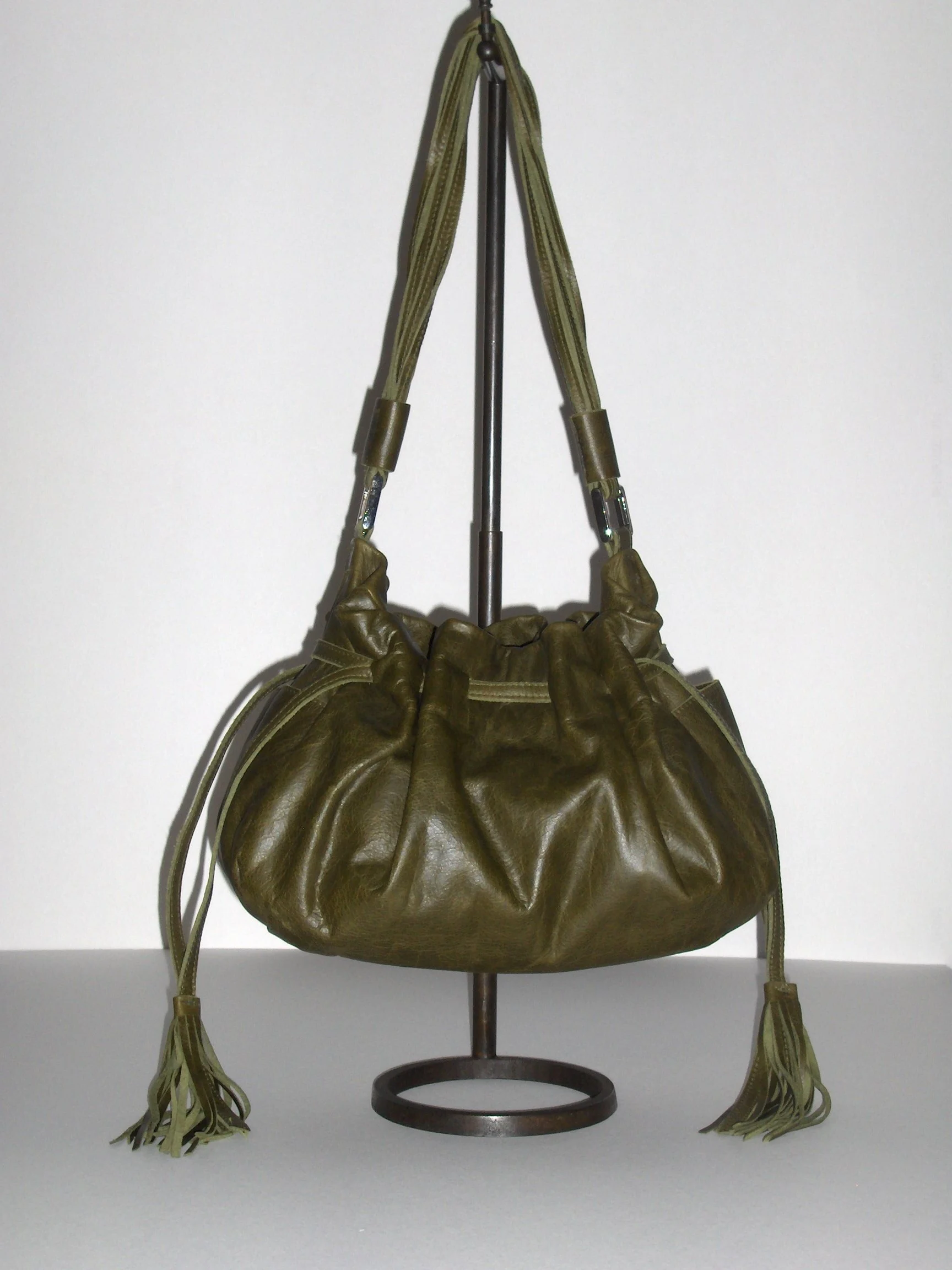Olive green leather handbag with tassel details hanging on a metal hook stand against a white background.