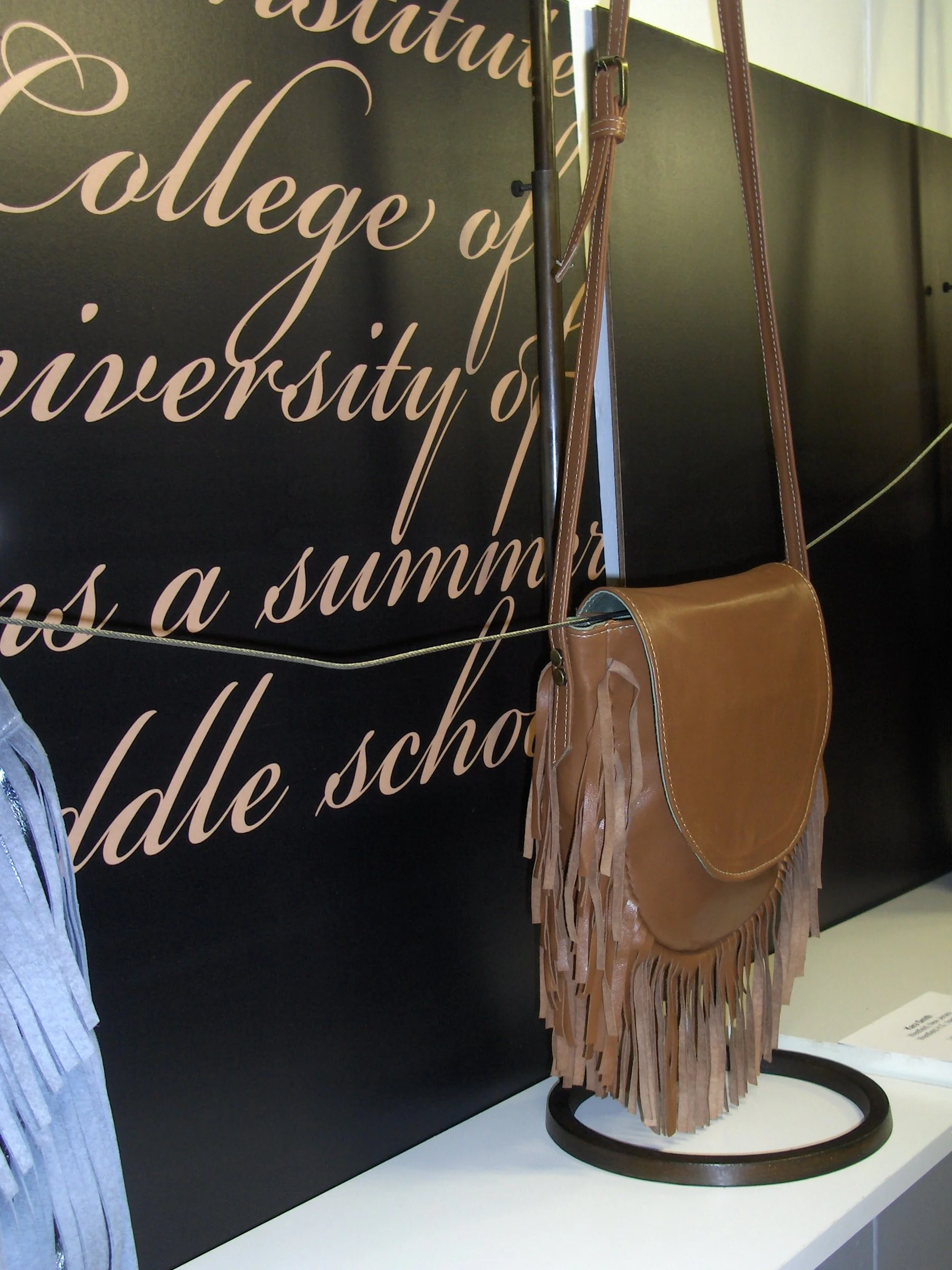 A tan fringe leather handbag with a long strap displayed on a stand against a black wall with cursive text in the background.