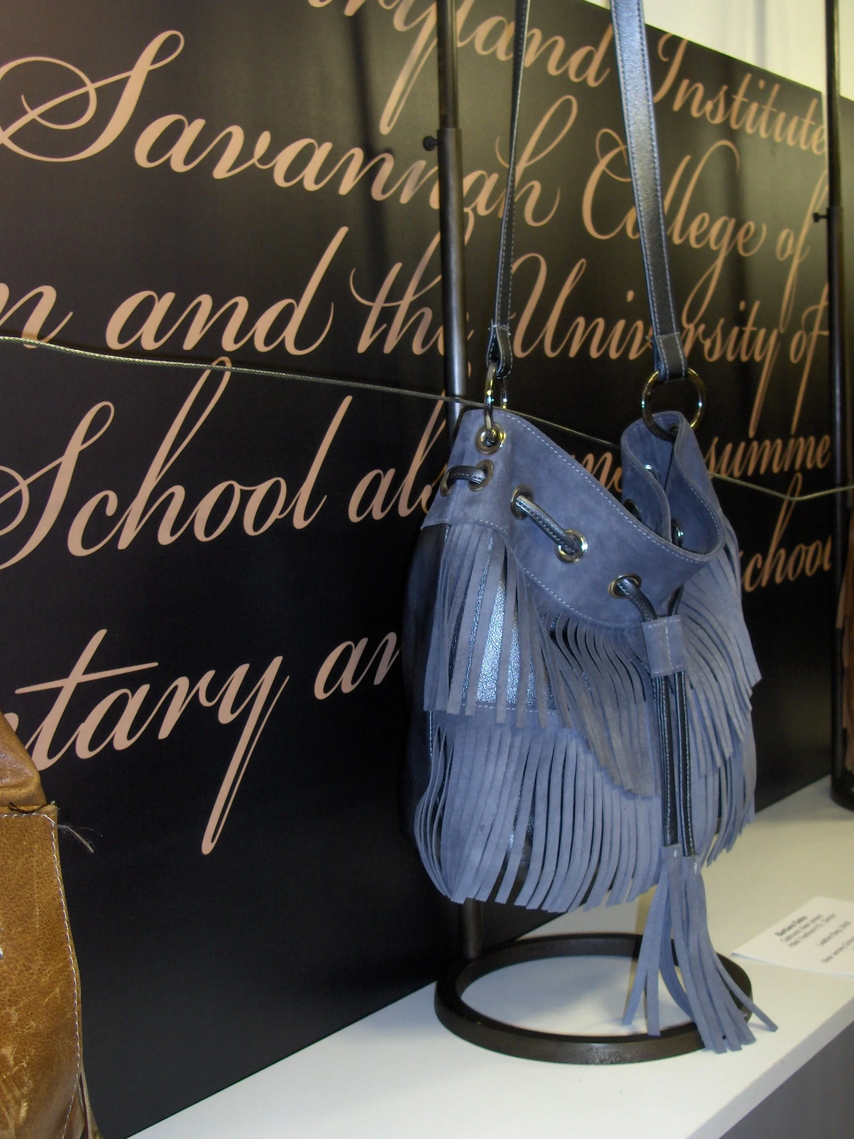 A fringed blue leather handbag with a zipper and grommet details, hanging on a display stand against a black background with pink cursive text.