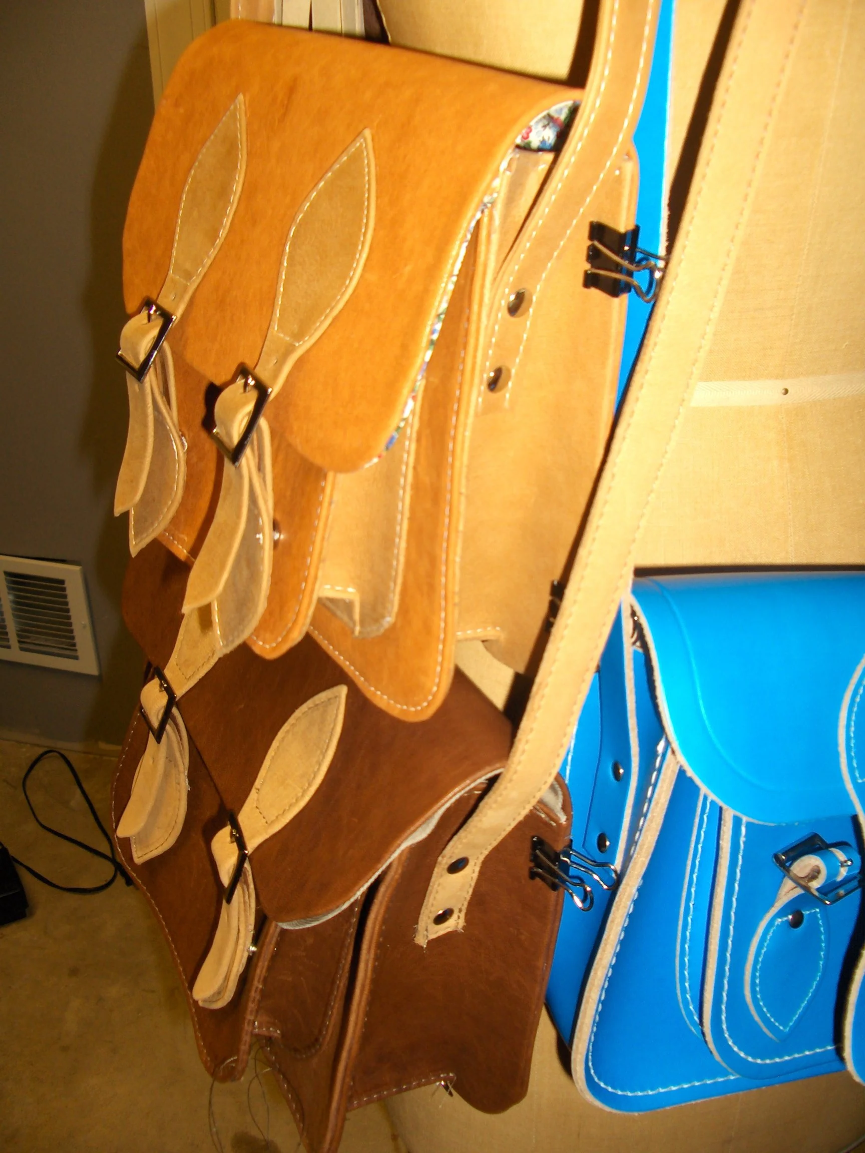 A display of three leather handbags with buckle straps, arranged on a surface. The handbags are in tan, brown, and blue colors.