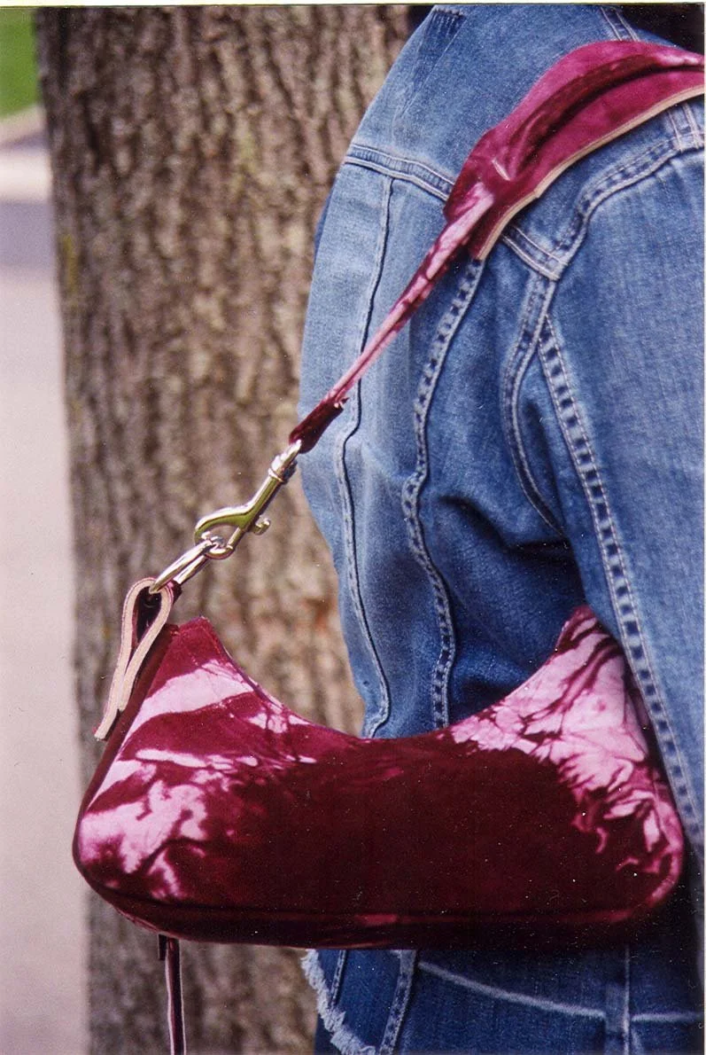 Person wearing blue jeans carrying a maroon and white tie-dye handbag on a shoulder strap, standing near a tree.