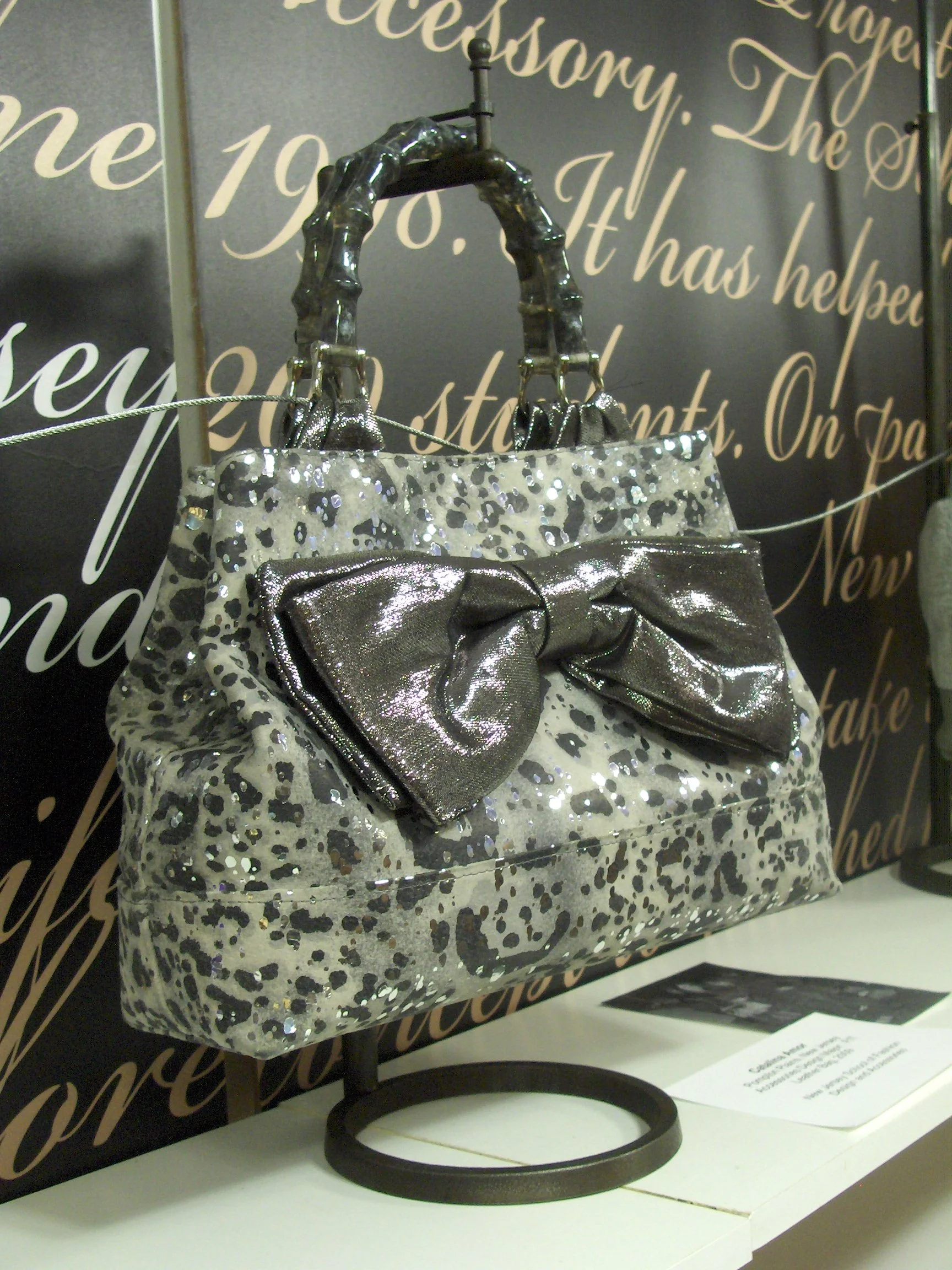 A boutique display features a silver and black leopard print handbag with a large metallic bow on the front, hanging on a black circular stand.