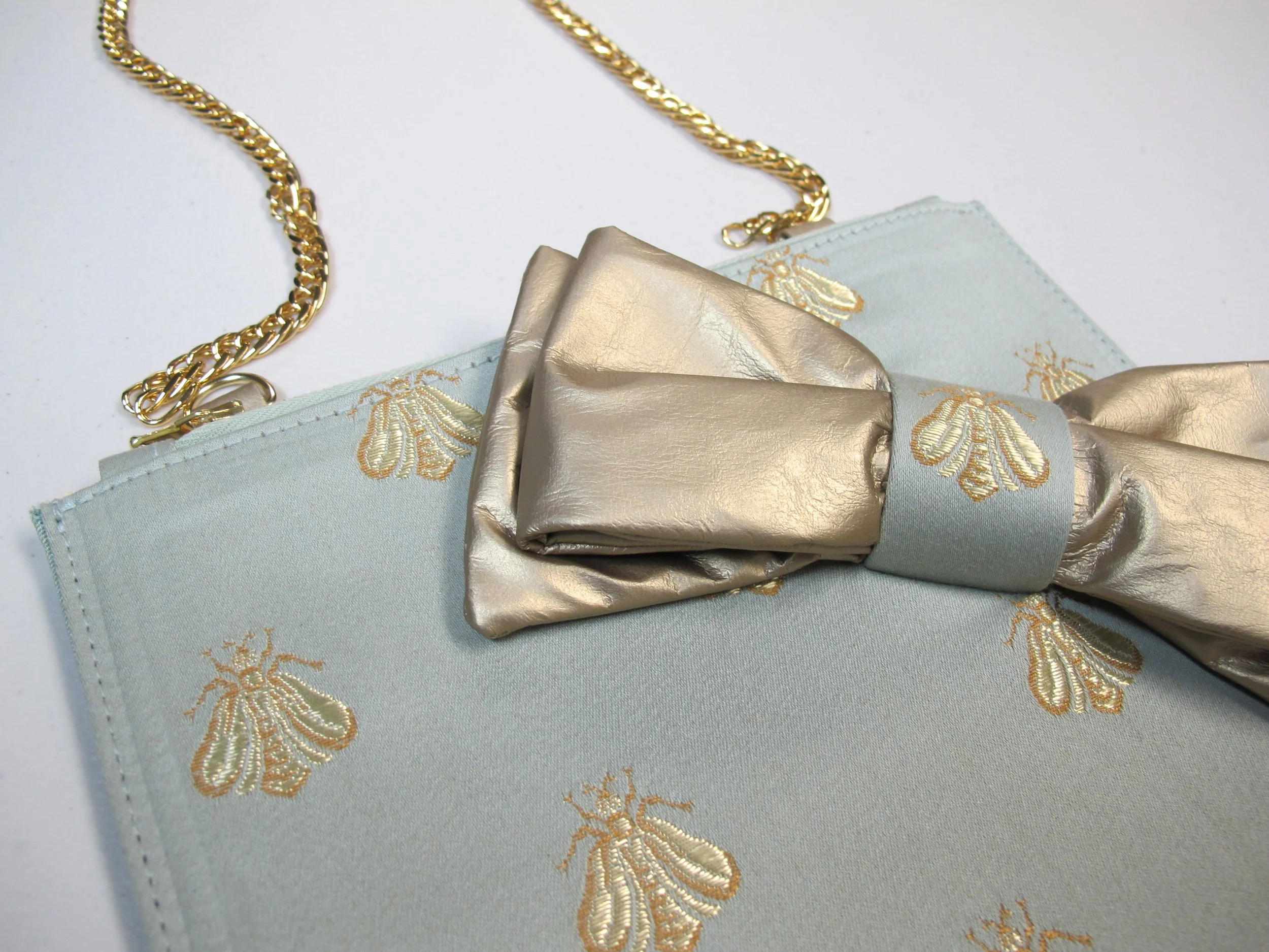 Close-up of a light blue clutch with gold embroidered bees, featuring a gold chain strap and a metallic gold bow with a light blue fabric band.