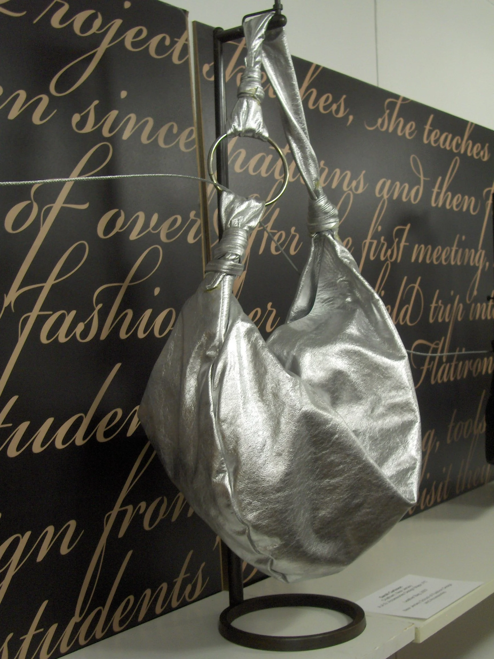 A metallic silver handbag hanging from a circular ring attached to a stand, with a background of large, stylish script writing on a dark wall.