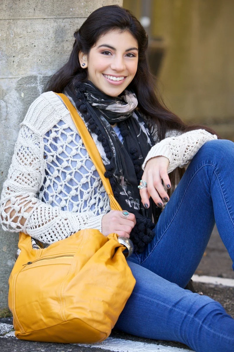 A young woman with long dark hair, wearing a white crochet sweater, black scarf, and blue jeans, sitting on the ground next to a stone wall, smiling at the camera, holding a yellow bag.