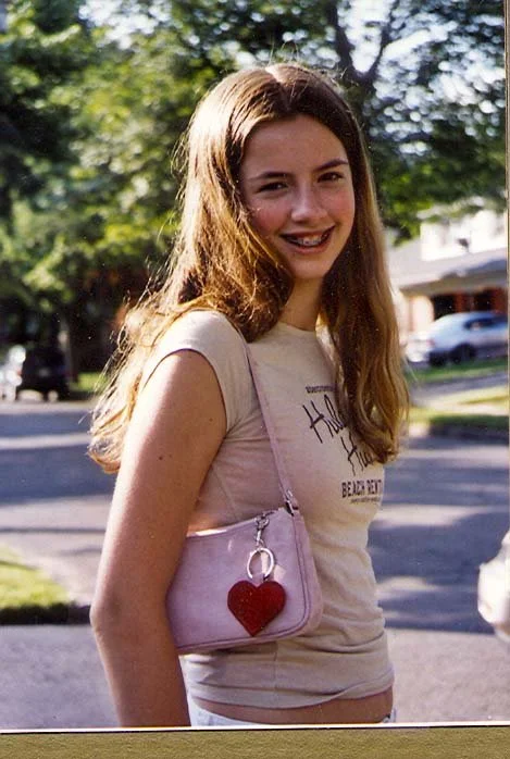 A young woman with long, wavy brown hair smiling outdoors, wearing a light-colored T-shirt with text, carrying a small pink purse with a red heart ornament, on a sunny day with trees and houses in the background.