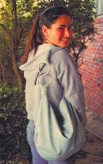 Young woman with long dark hair smiling, wearing a light gray hoodie, standing outdoors near a brick wall and trees.