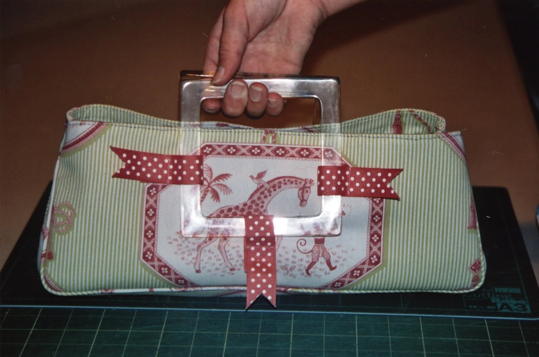 A vintage-style bag with a giraffe design, red polka dot ribbon handle, and a clear window on the front being held by a person's hand.
