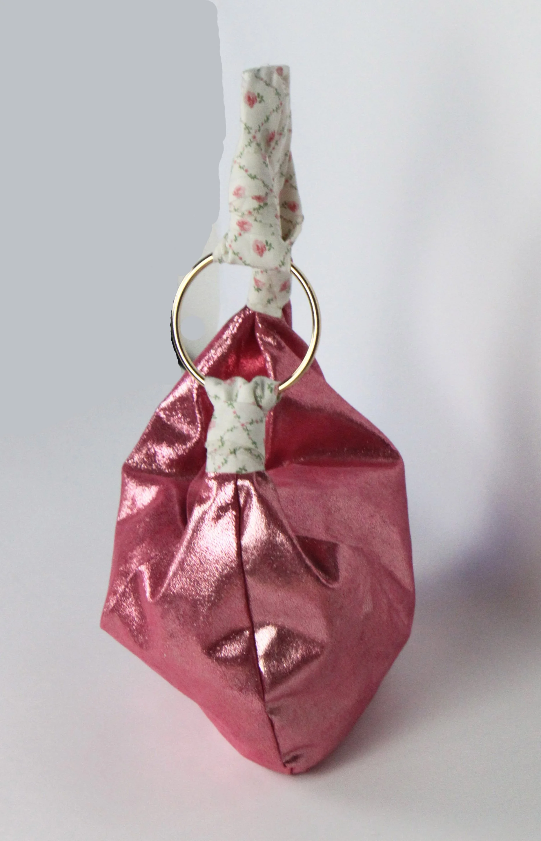 A pink metallic gift bag with a floral fabric handle and a decorative ring. The bag is folded at the top, and the handle is tied with matching floral fabric.